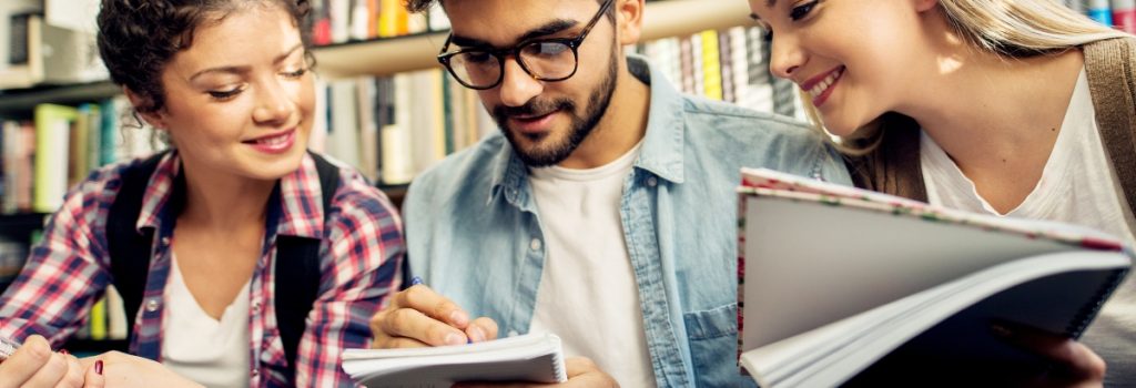 Portrait of a young joyful male student learning on a library desk on which two female students are studying in a background.